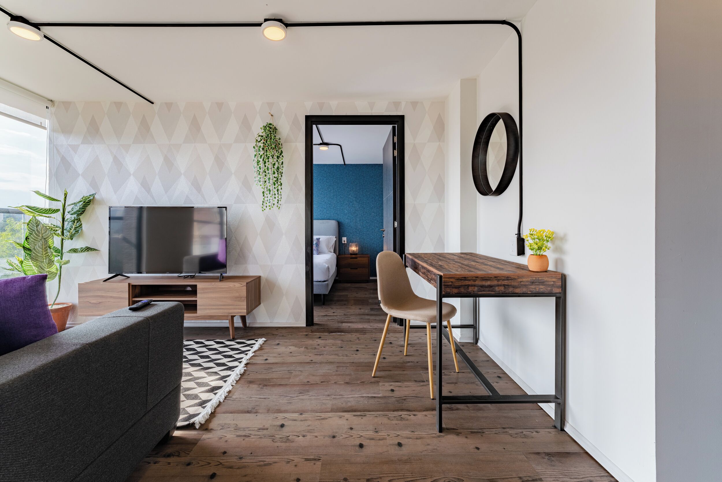 Un espacio habitable moderno con televisor, sofá, mesa de comedor y vista hacia un dormitorio.
