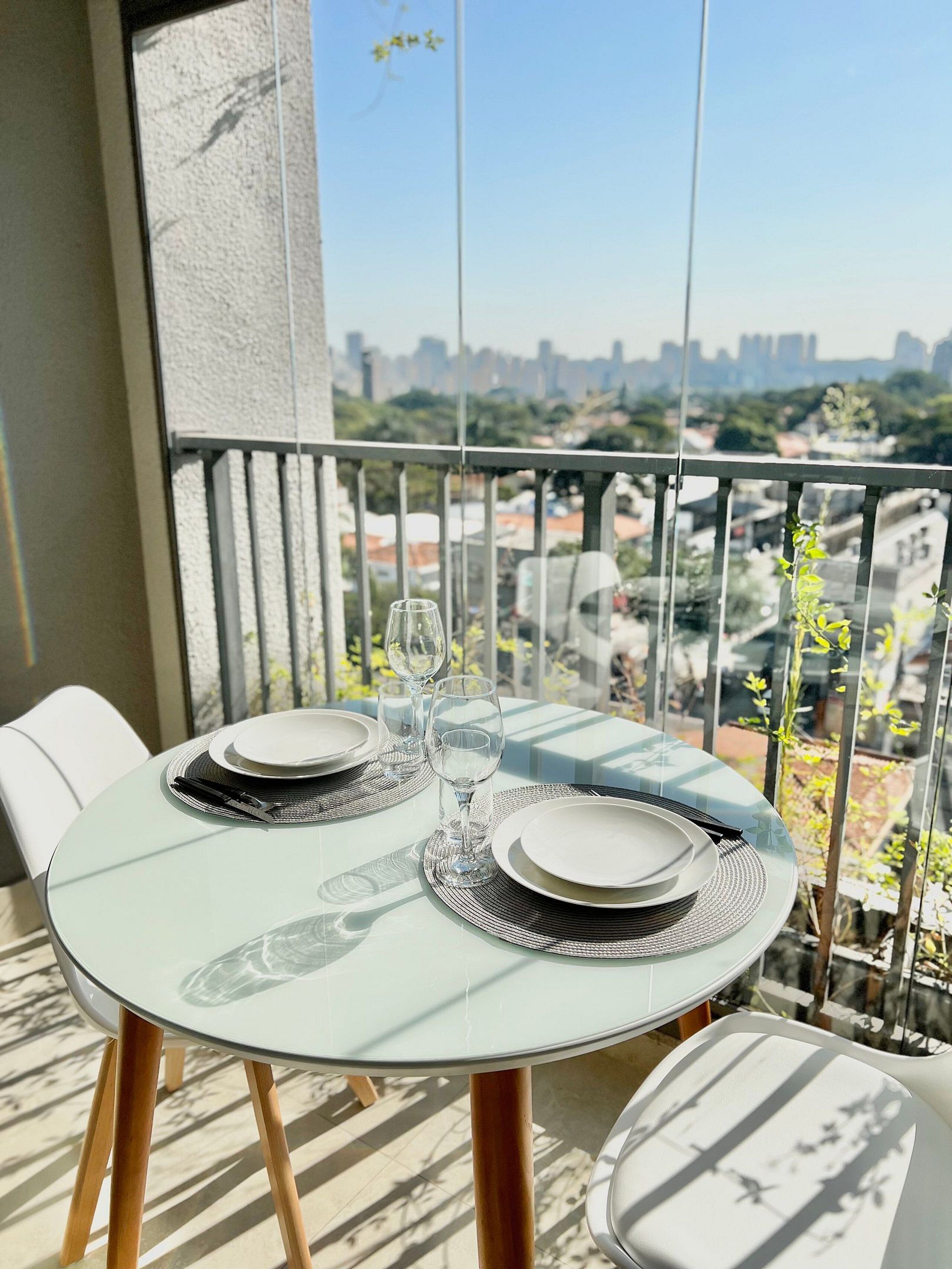 A round table with two plates, two wine glasses, and cutlery on a balcony with a city view.