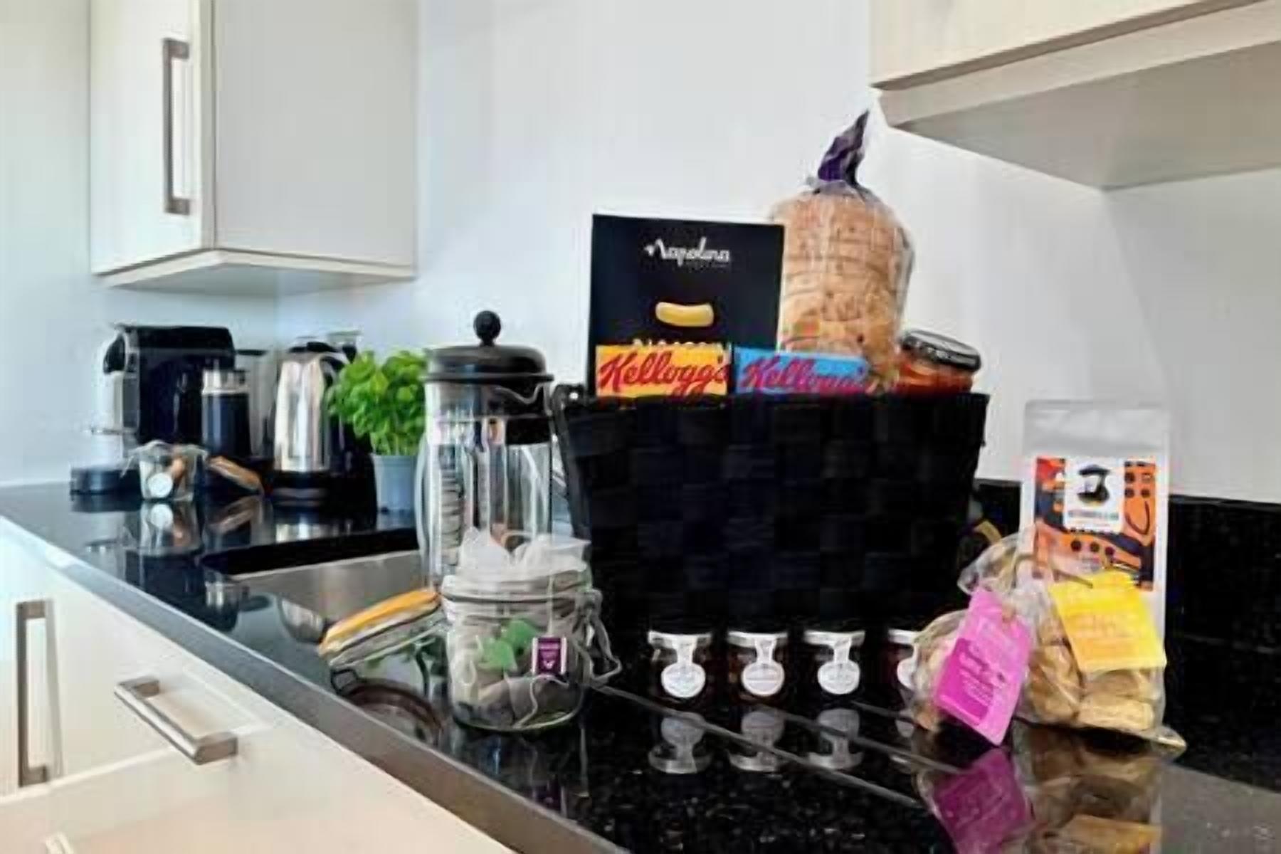 Una encimera de cocina con una cafetera, un hervidor, un tarro de pasta, un paquete de pan y una caja de cereales Kellogg's.