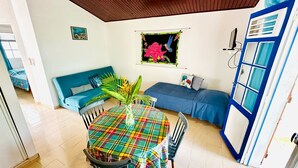 Une pièce chaleureuse avec un canapé bleu, une table aux couleurs vives et une fenêtre ornée d’un tableau représentant des fleurs.