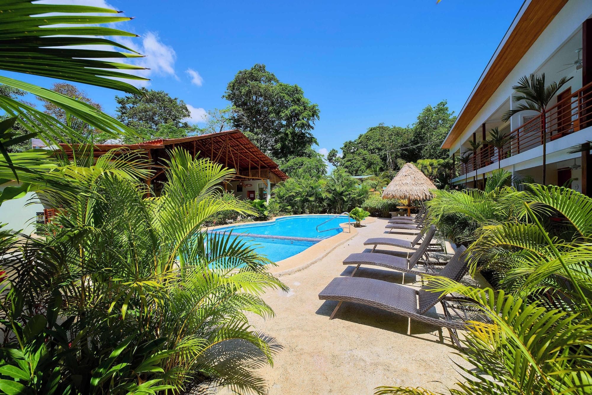 Zona de piscina con sillones de descanso, una sombrilla de paja y vegetación tropical.