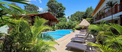 Zona de piscina con sillones de descanso, una sombrilla de paja y vegetación tropical.