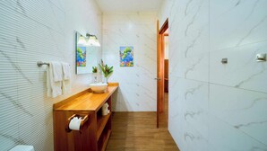 A modern bathroom with a wooden vanity, white marble tiles, a large mirror, and a painting on the wall. - Olinca Boutique Hotel (Cahuita)