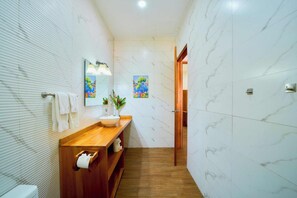 A modern bathroom with a wooden vanity, white marble tiles, a large mirror, and a painting on the wall. - Olinca Boutique Hotel (Cahuita)