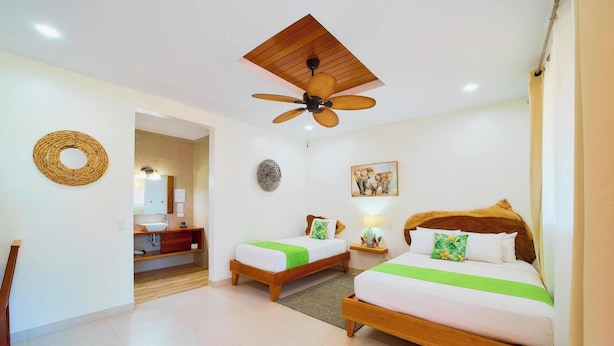 A hotel room with two beds, a ceiling fan, a wooden headboard, a bathroom with a sink and mirror, and a woven wall decoration. - Olinca Boutique Hotel (Cahuita)