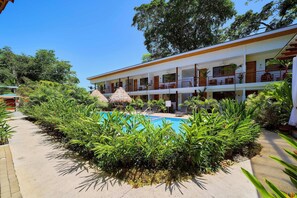 Hotel com piscina rodeada por plantas tropicais, estrutura com telhado de palha e edifício com várias andares e varandas.