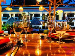 Mesa posta para refeição com vários copos de vinho e talheres, um guarda-sol de palha, e vista para um edifício com janelas e plantas ao fundo.