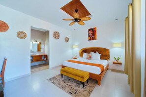 Quarto moderno com cabeceira de madeira, uma cama grande, um banco amarelo, um ventilador de teto e um banheiro visível por uma porta aberta.