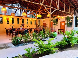 Un área de comedor al aire libre con mesas y sillas de madera, rodeada de plantas en macetas y una estructura de techo de paja.