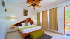 A bedroom with a wooden headboard, a bed with white and green bedding, a green ottoman, a ceiling fan, and a view of greenery through the window. - Olinca Boutique Hotel (Cahuita)