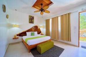 A bedroom with a wooden headboard, a bed with white and green bedding, a green ottoman, a ceiling fan, and a view of greenery through the window. - Olinca Boutique Hotel (Cahuita)