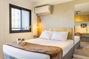 A hotel room with a bed, a window, an air conditioner, and a nightstand with a lamp.