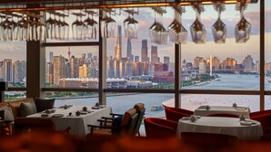 A dining area with tables and chairs, large windows with a city skyline view, and hanging light fixtures. - Thompson Shanghai Expo, by Hyatt (Shanghai)