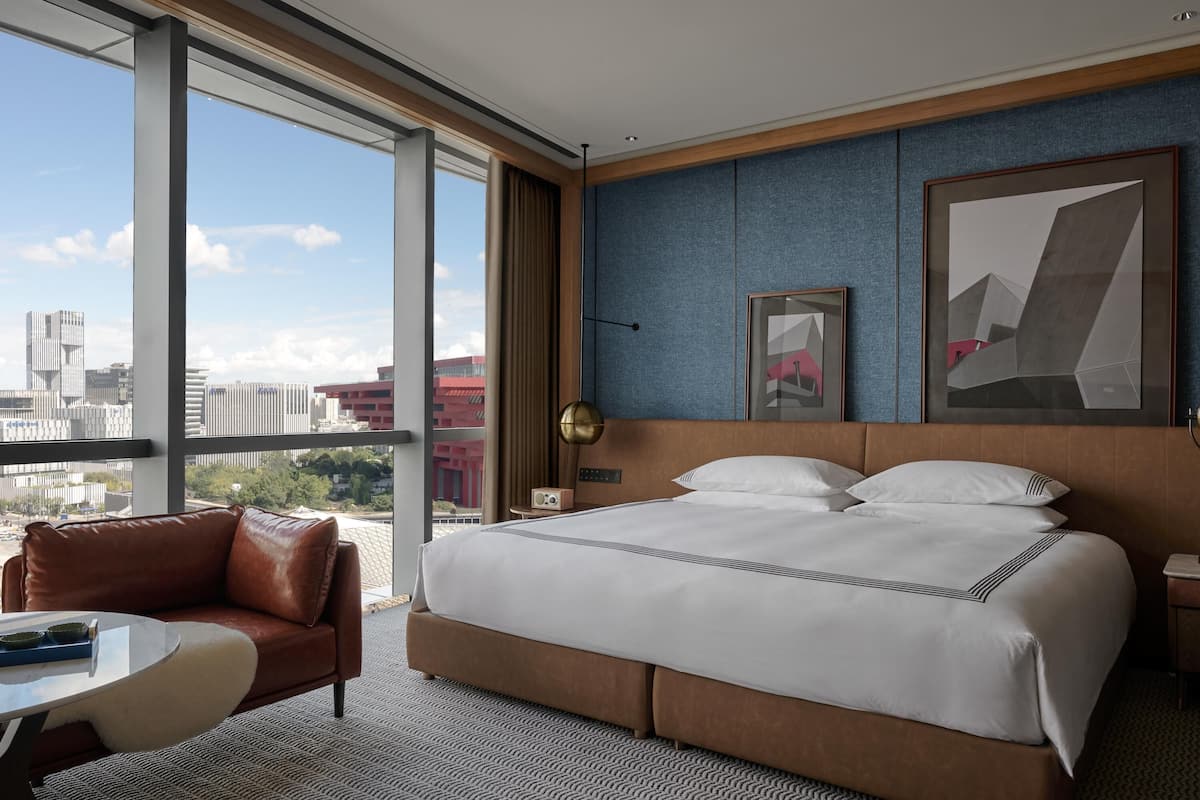 A hotel room with a large bed, a leather armchair, a small table, and a city view.