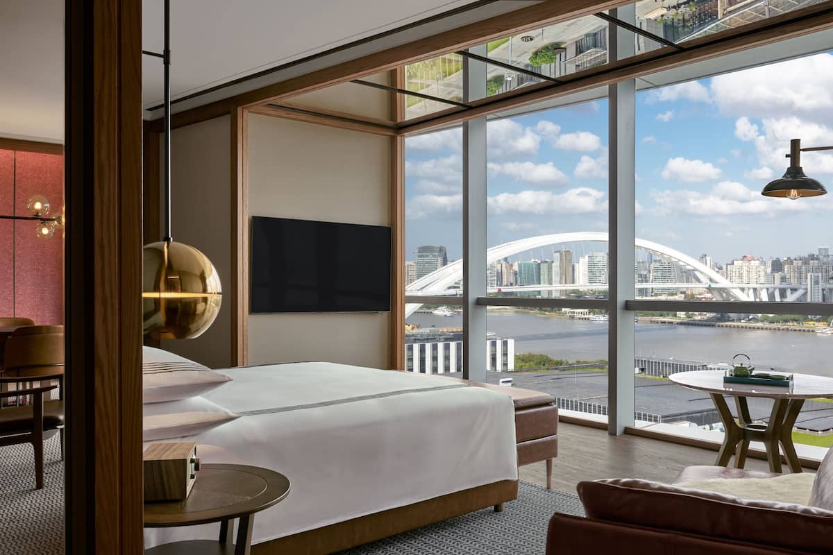 A modern hotel room with a large bed, a TV, a dining table with a kettle, and a view of a cityscape and river.