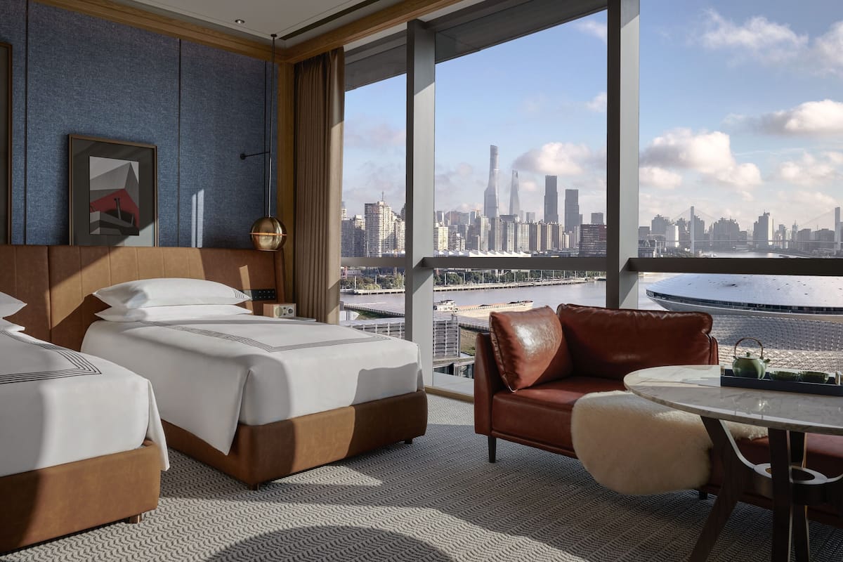 A hotel room with two beds, a leather sofa, a round table, and a large window offering a city skyline view.