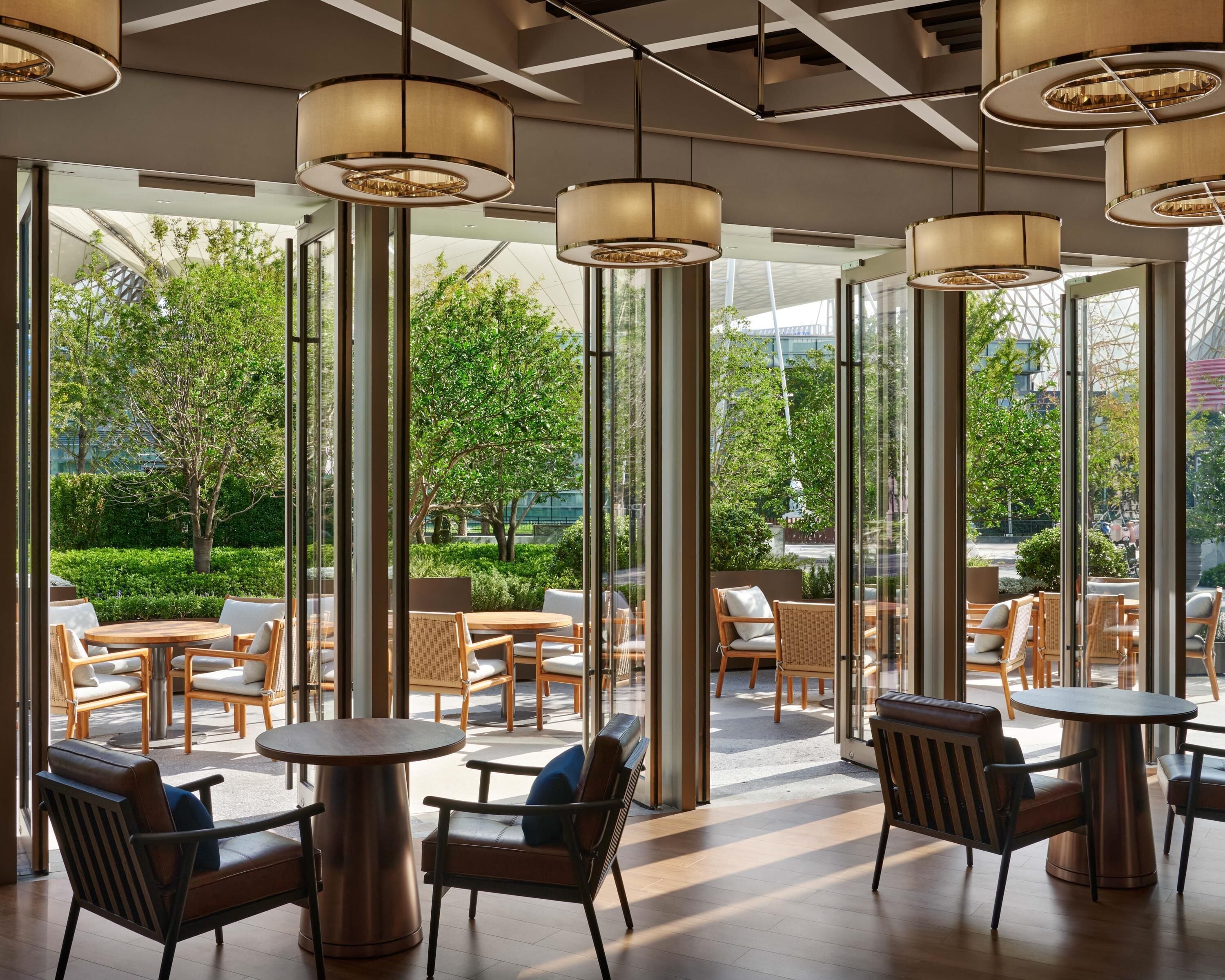 A modern indoor seating area with round tables and chairs, large windows, and hanging pendant lights.