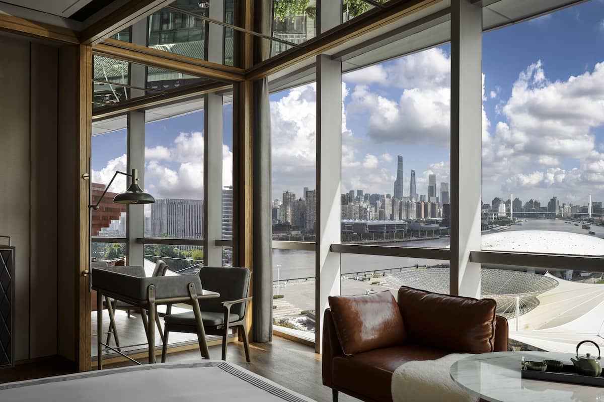 A modern hotel room with a large window offering a city view, a leather sofa, a small table with a teapot, and a desk with a lamp.