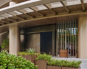 A modern building entrance with a glass door, a wooden bench, and planters with greenery. - Thompson Shanghai Expo, by Hyatt (Shanghai)