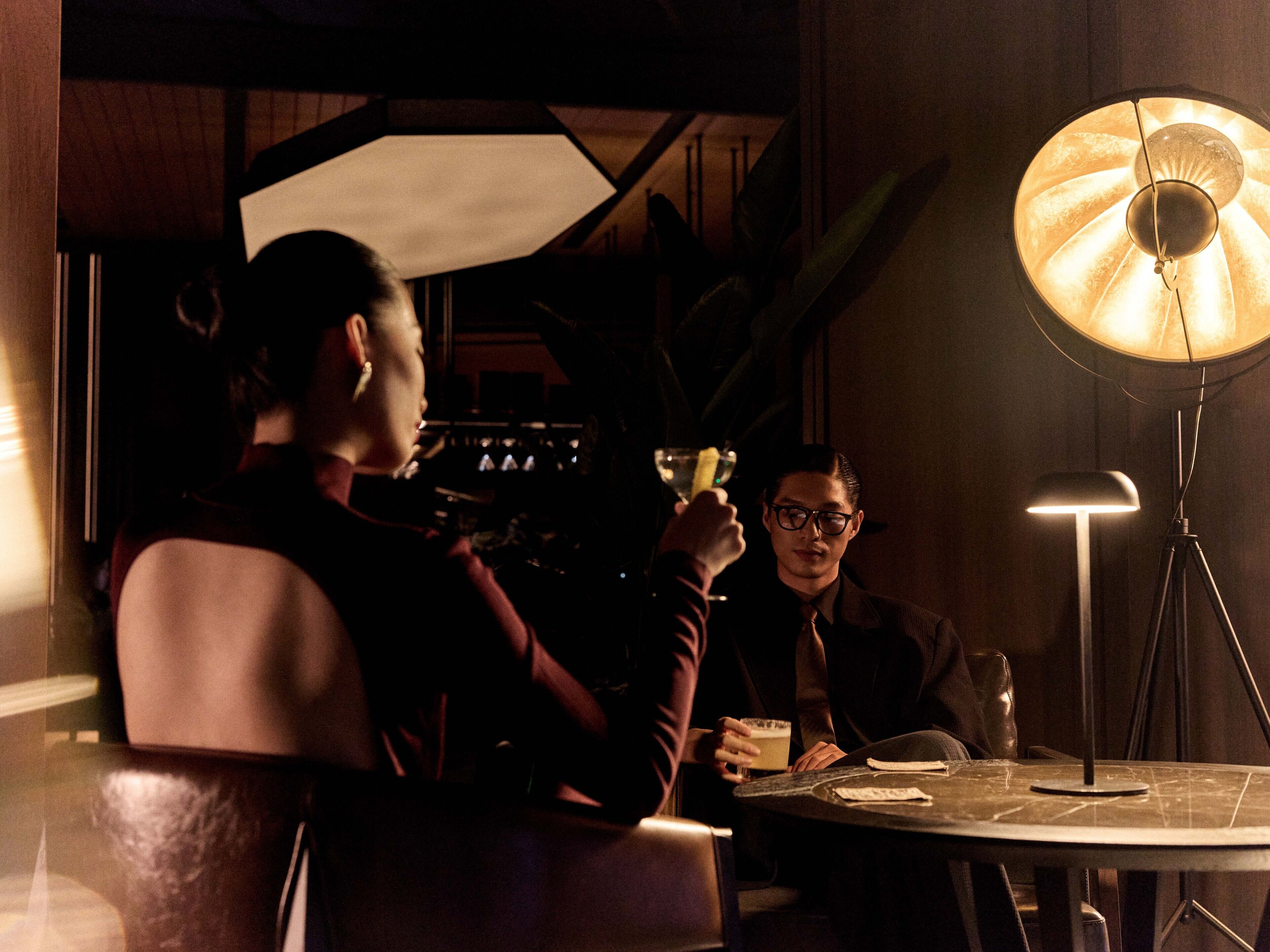 Two people seated at a round table in a dimly lit room with a large floor lamp.