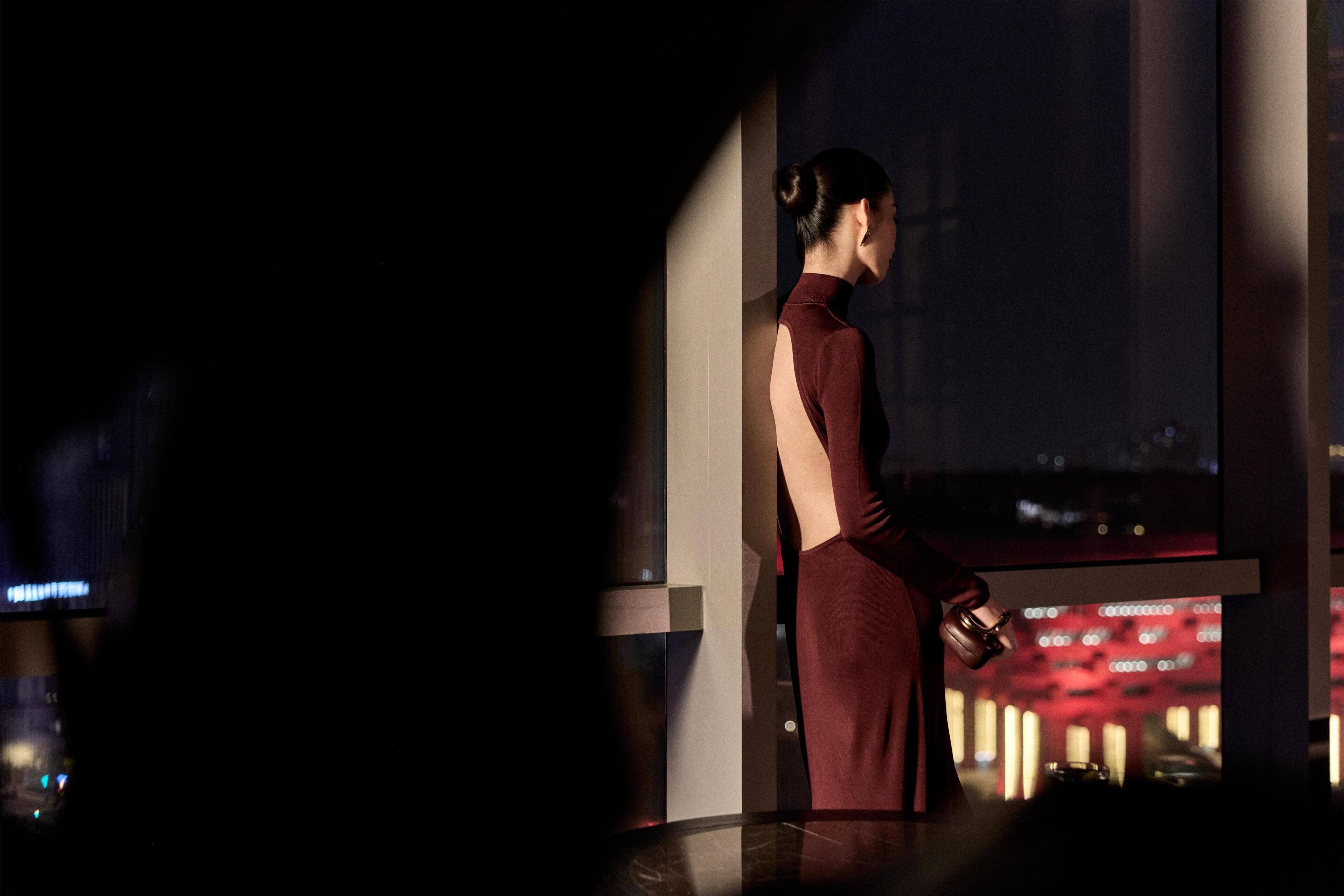A person in a red dress standing by a window with a cityscape view.