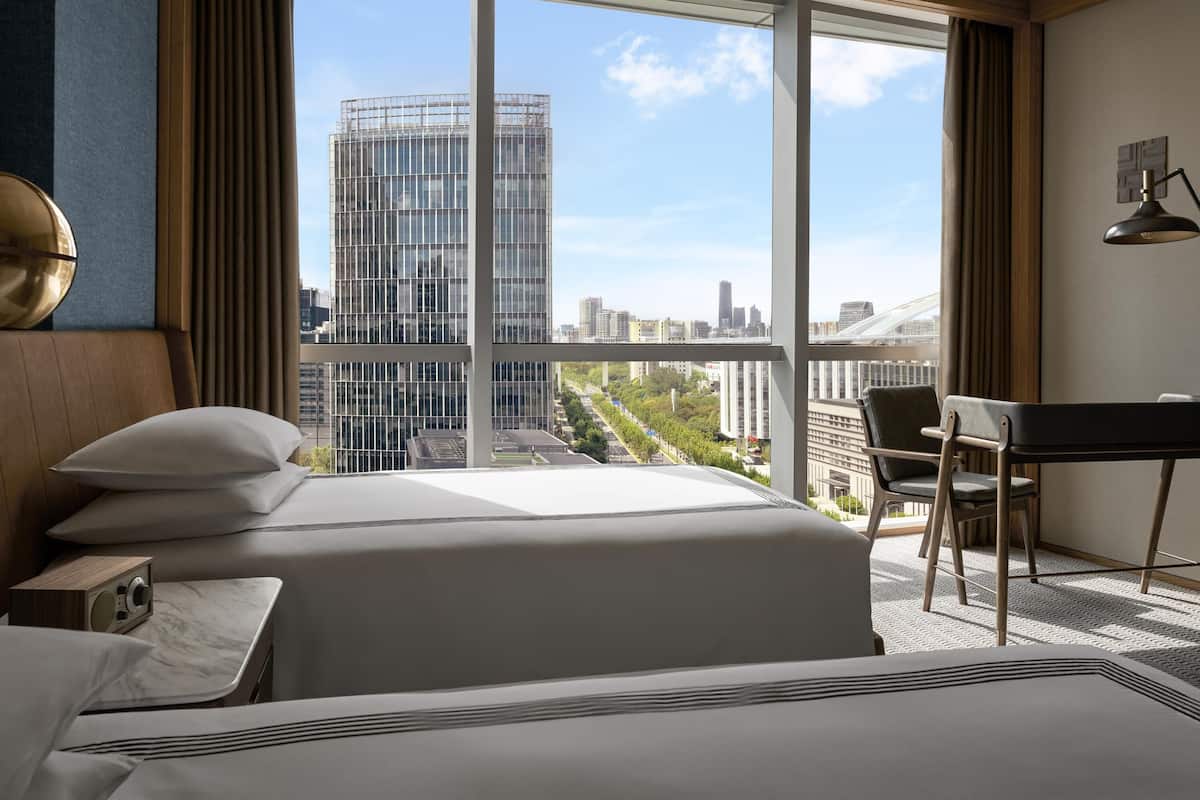 A hotel room with a large bed, a desk, a chair, and a view of a cityscape through large windows.