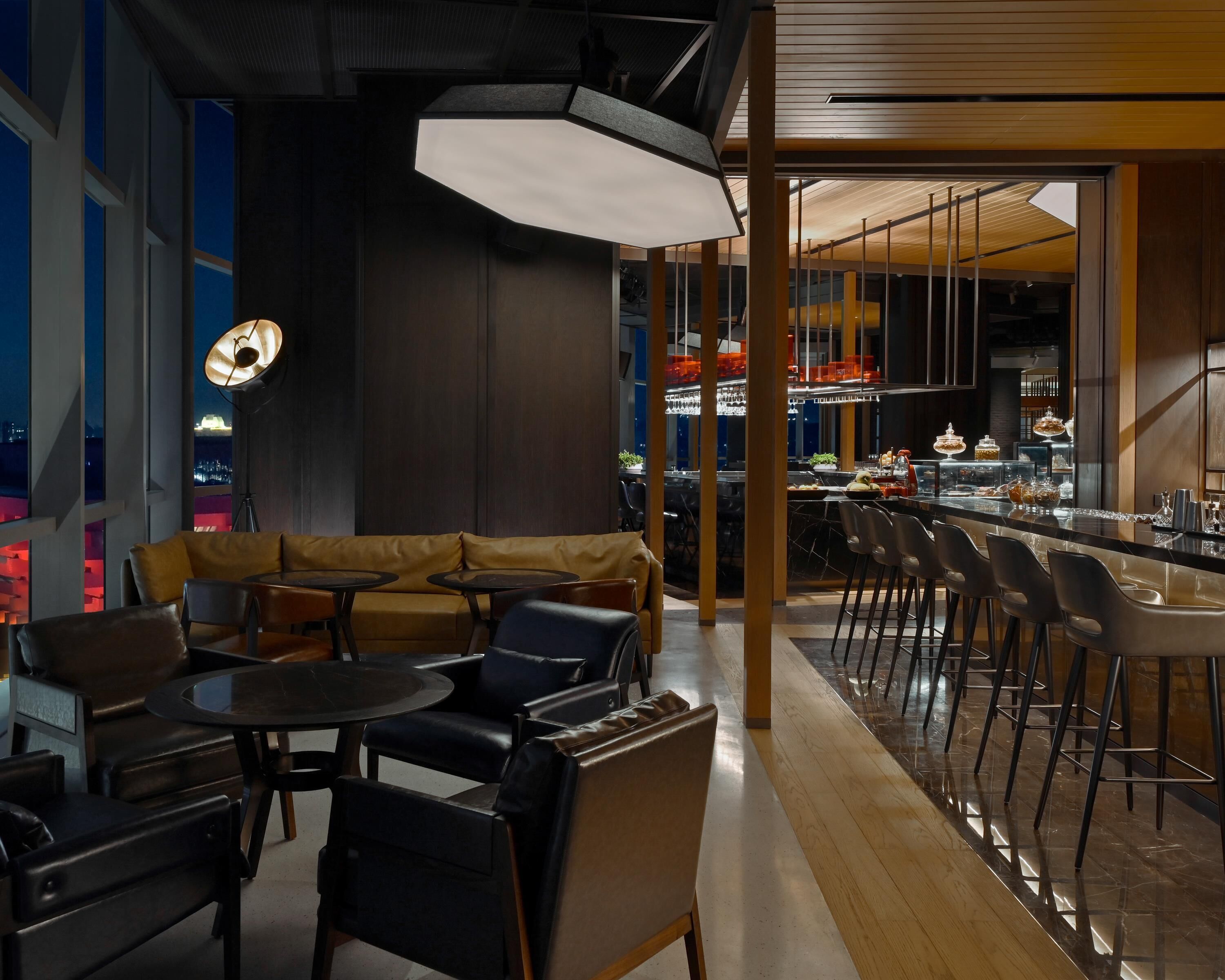A modern bar area with leather seating, a bar counter, and a view of the city at night.