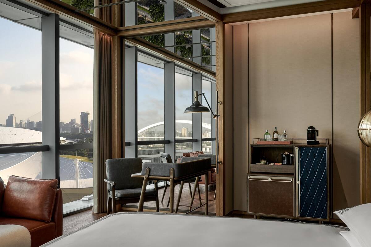 A modern hotel room with a large bed, a desk with chairs, a minibar, and a view of a cityscape through large windows.