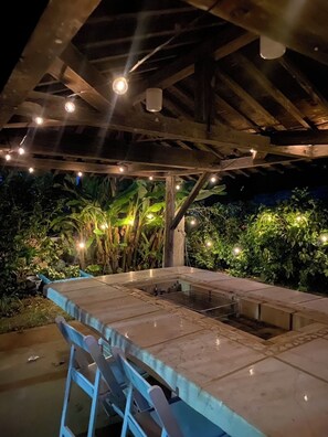 Un patio al aire libre con una mesa y sillas, rodeado de abundante vegetación y luces colgantes.