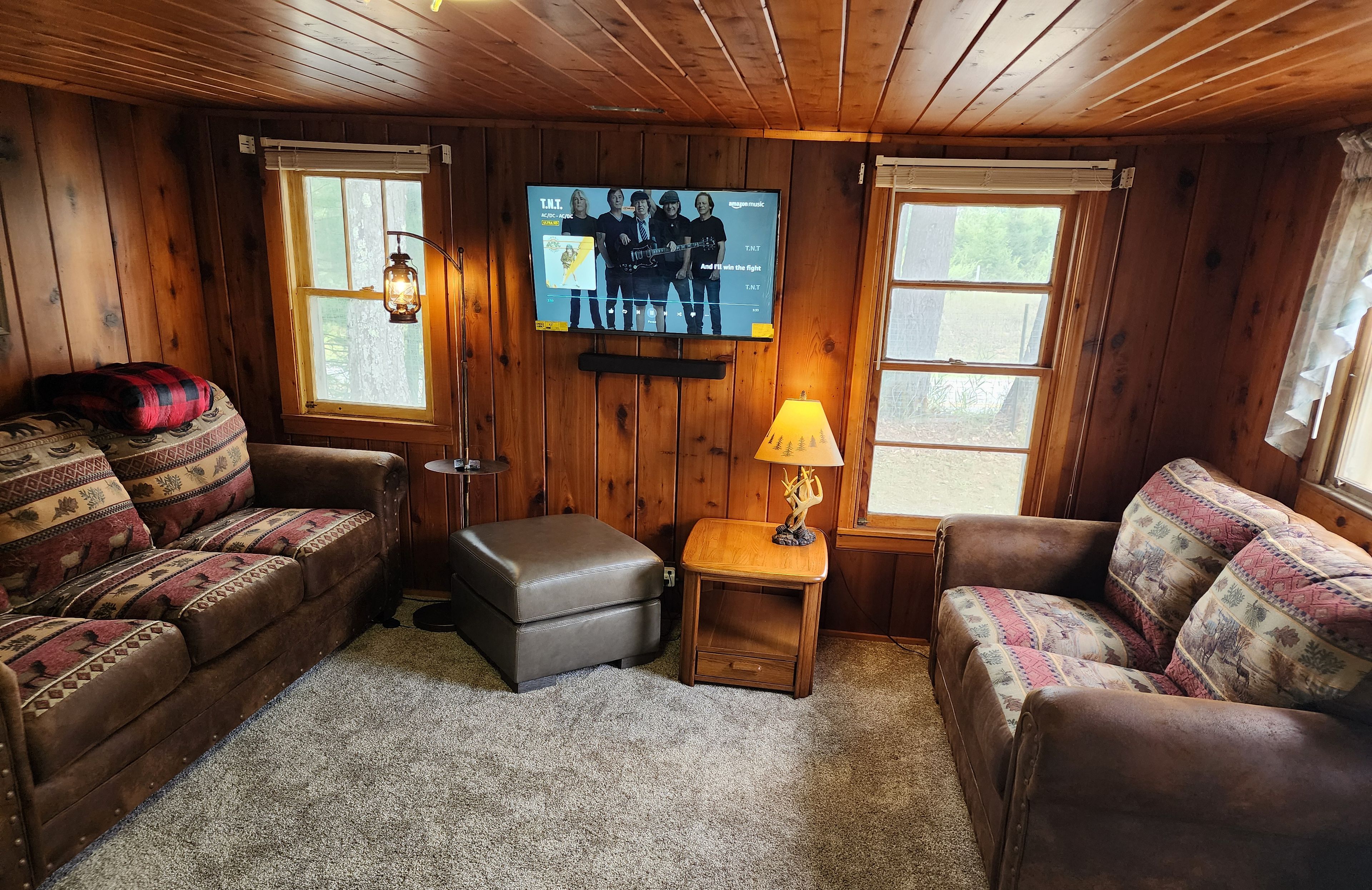 Acogedora habitación tipo cabaña con paredes de madera, dos sofás, una mesita con una lámpara, un televisor en la pared y una ventana.