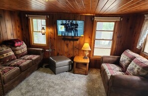 A cozy cabin room with wooden walls, two sofas, a small table with a lamp, a TV on the wall, and a window.