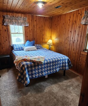 A cozy bedroom with wooden walls, a large bed with blue and white checkered bedding, a window with a view of the outdoors, and a nightstand with a lamp.