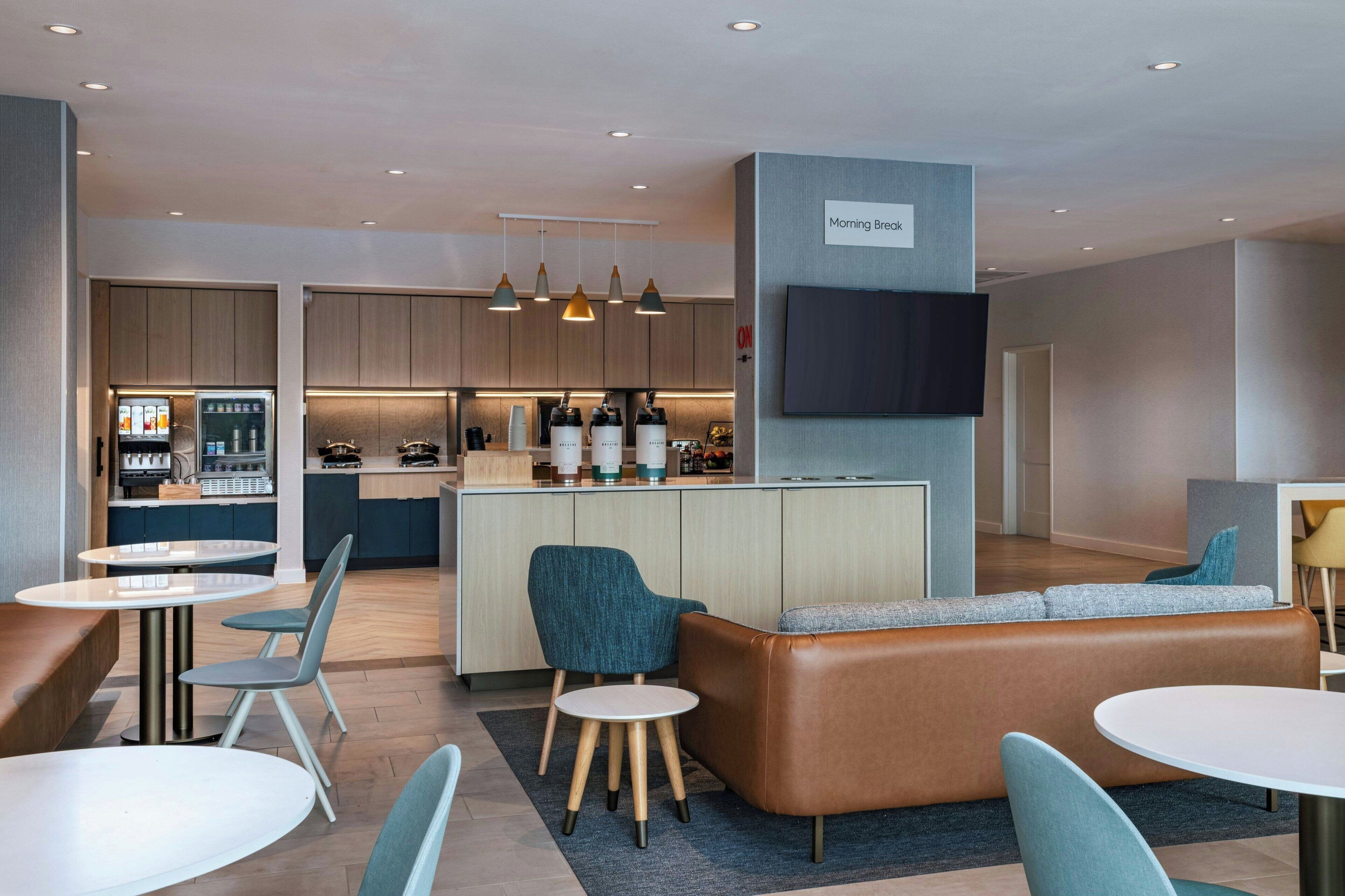 Um hall de hotel moderno com área de bar central, espaços de estar e cozinha ao fundo.
