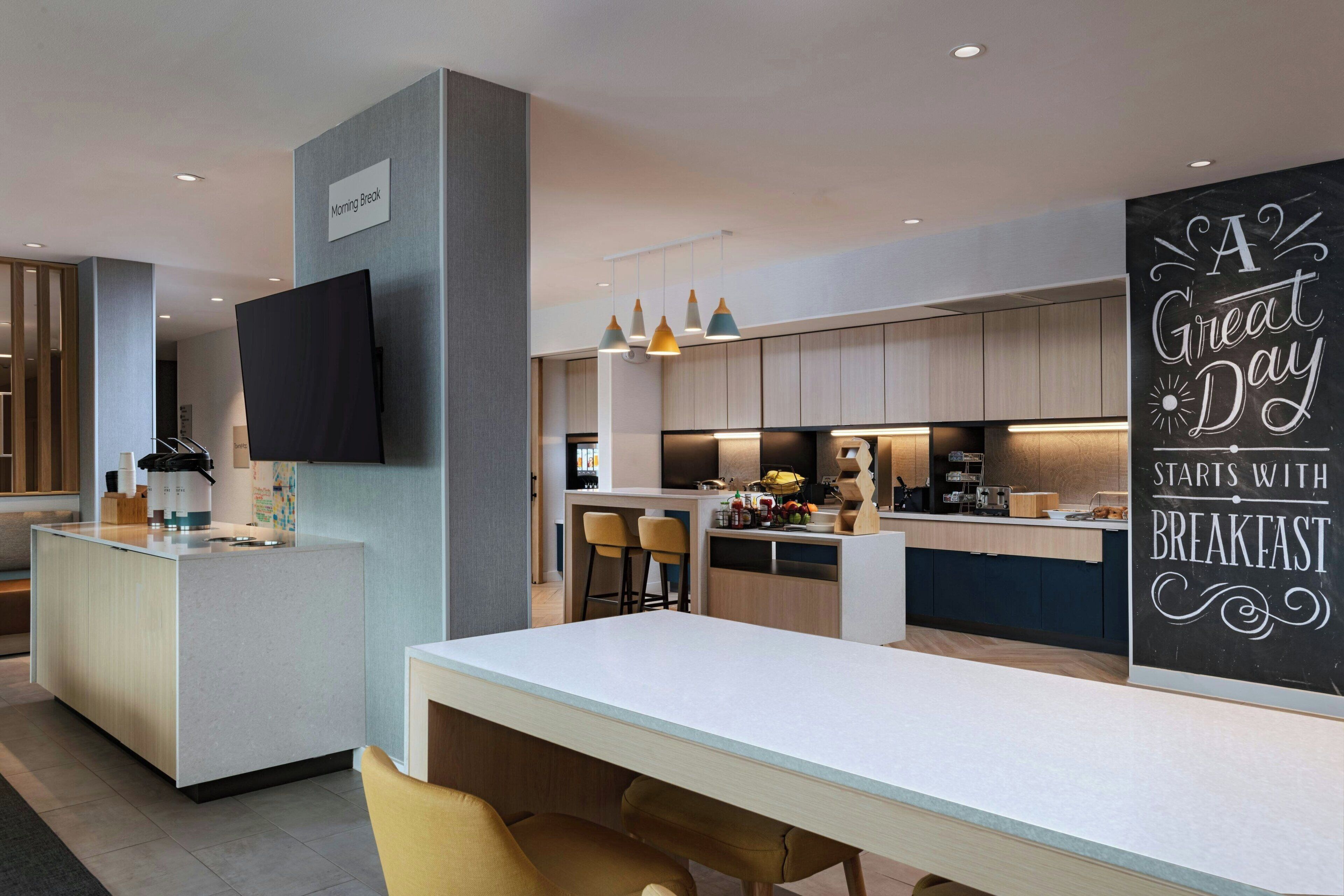 Cocina moderna con barra desayunadora, televisor de pantalla plana, lámparas colgantes y un cartel que dice 'A Great Day Starts with Breakfast'.