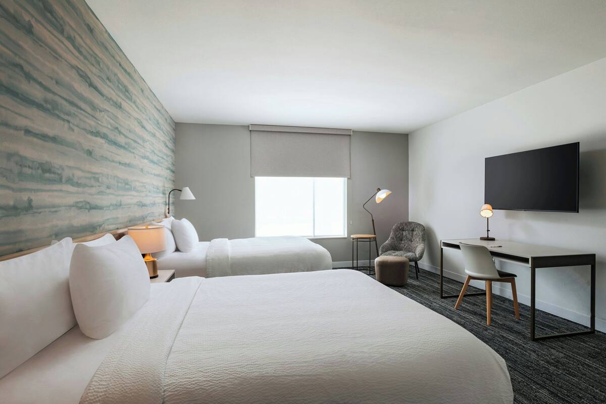 A hotel room with two beds, a desk, a chair, a TV, and a window with a view.