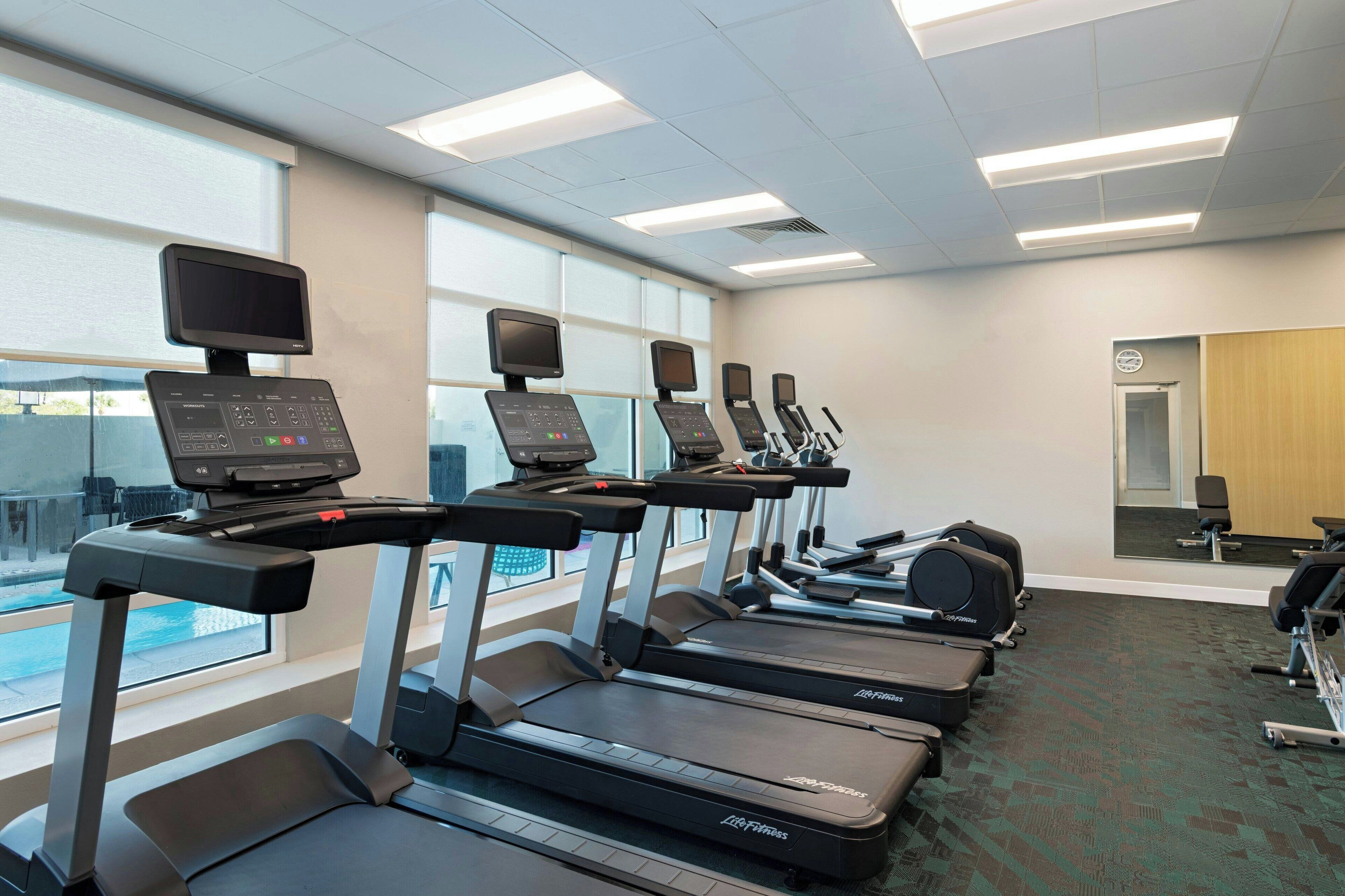A row of treadmills with digital screens, a large window, and a carpeted floor.