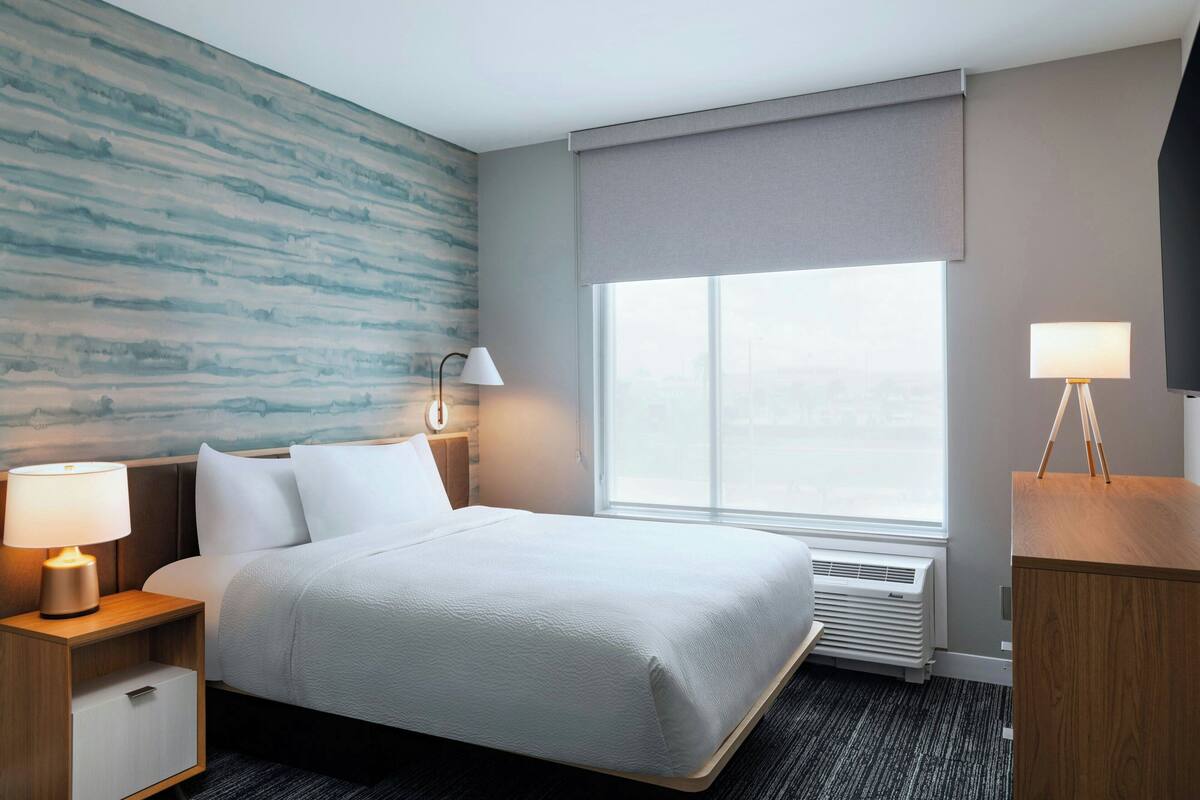 A hotel room with a large bed, a nightstand with a lamp, a bedside table with a lamp, a window with a view, and a wall with a textured design.