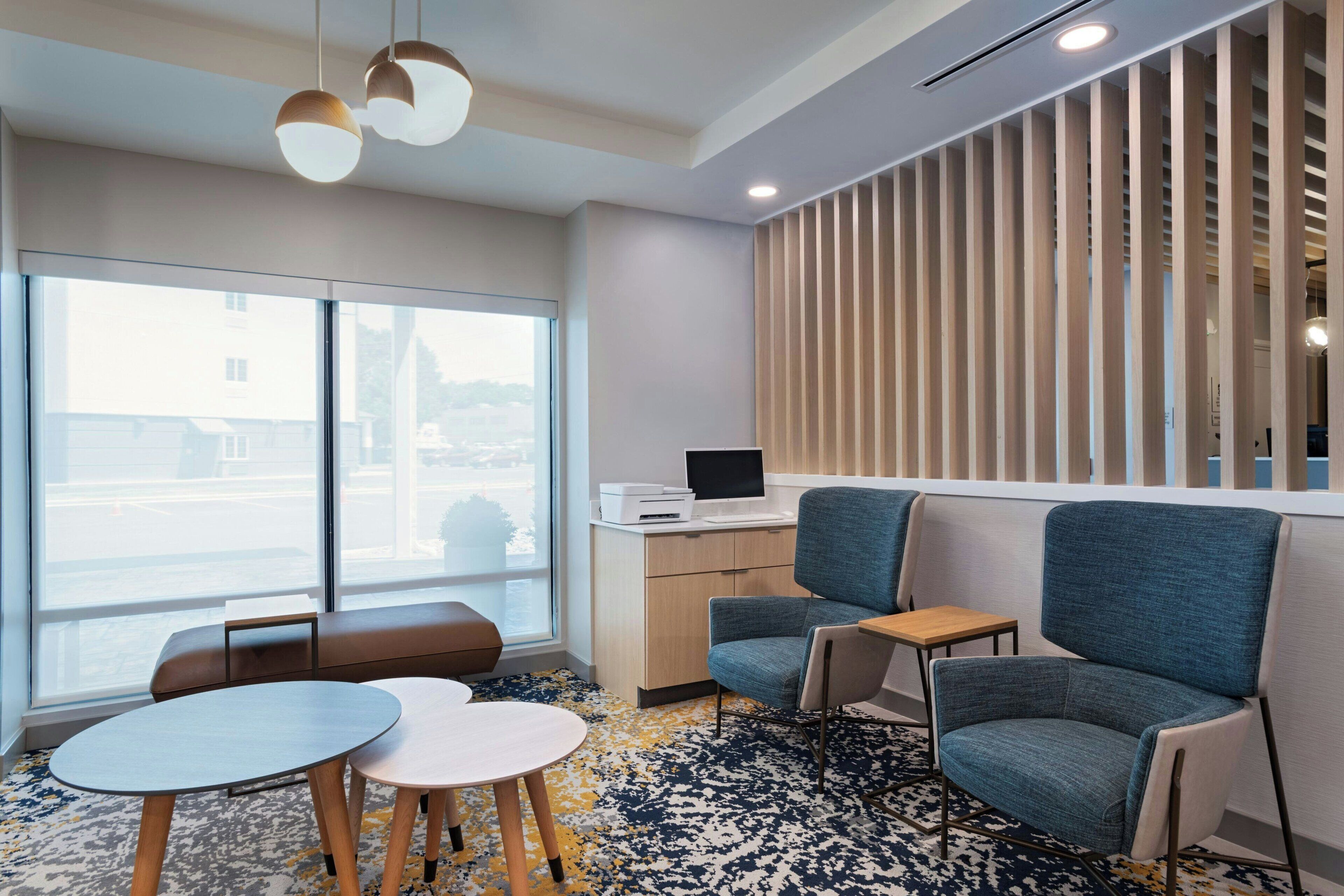 Une chambre d’hôtel moderne dotée d’une grande fenêtre, d’un coin salon avec un canapé et deux fauteuils, d’une petite table et d’un bureau avec un ordinateur.