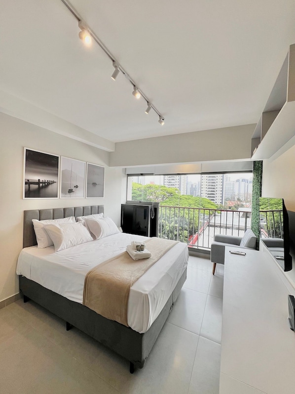 A modern bedroom with a large bed, a desk, a TV, and a balcony with a city view. - La Vida (São Paulo)