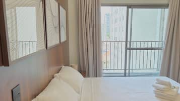 A hotel room with a large bed, a view of the city through a balcony, and a modern design with wooden paneling and artwork on the walls.