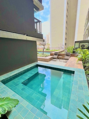 A swimming pool with clear blue water, surrounded by a tiled deck and some greenery. - La Vida (São Paulo)