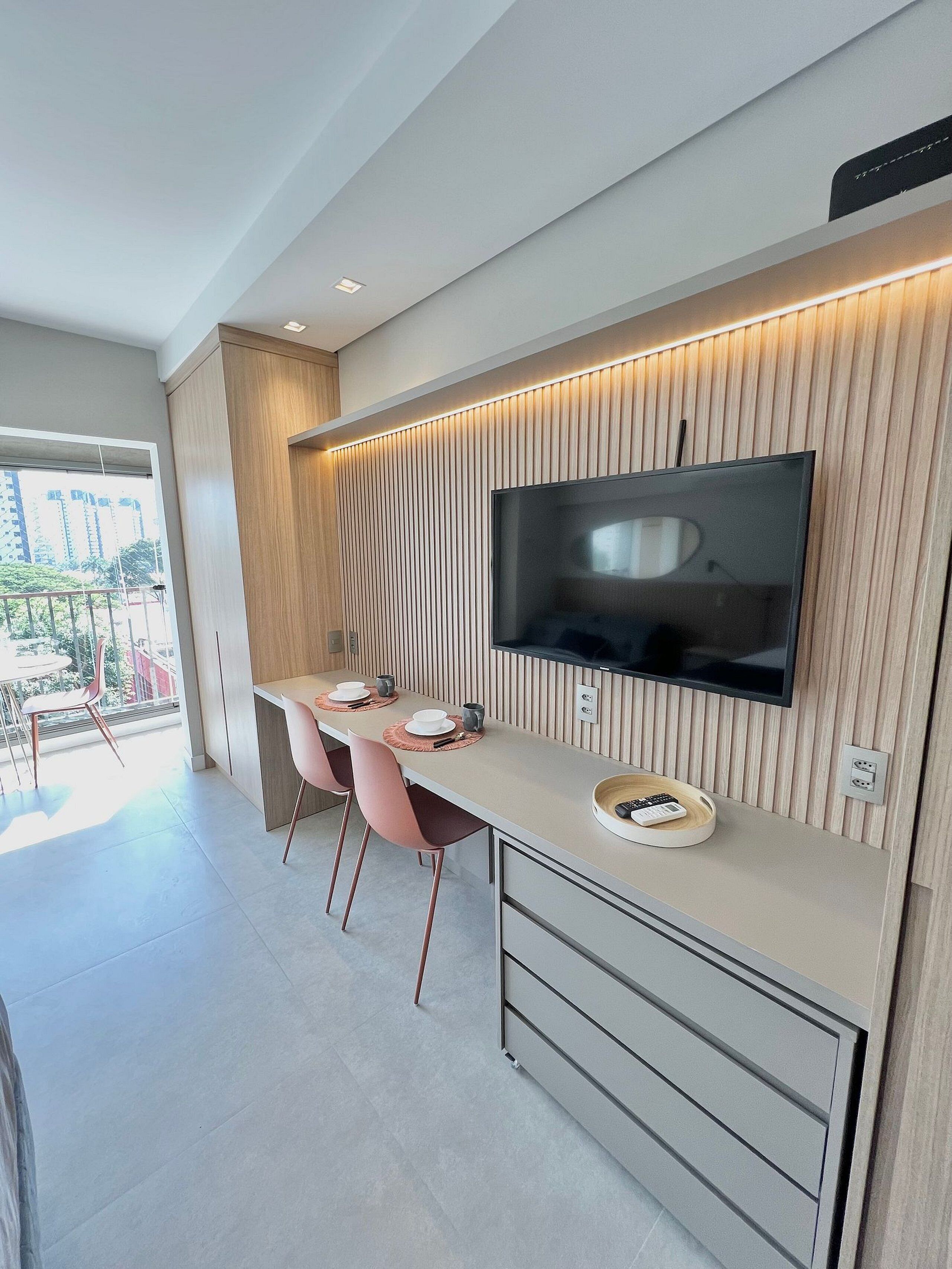 A modern kitchen with a built-in TV, wooden panel wall, and a dining area with pink chairs.