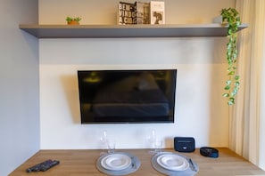 A flat-screen TV mounted on a wall above a wooden table with two plates, two glasses, and a remote control.