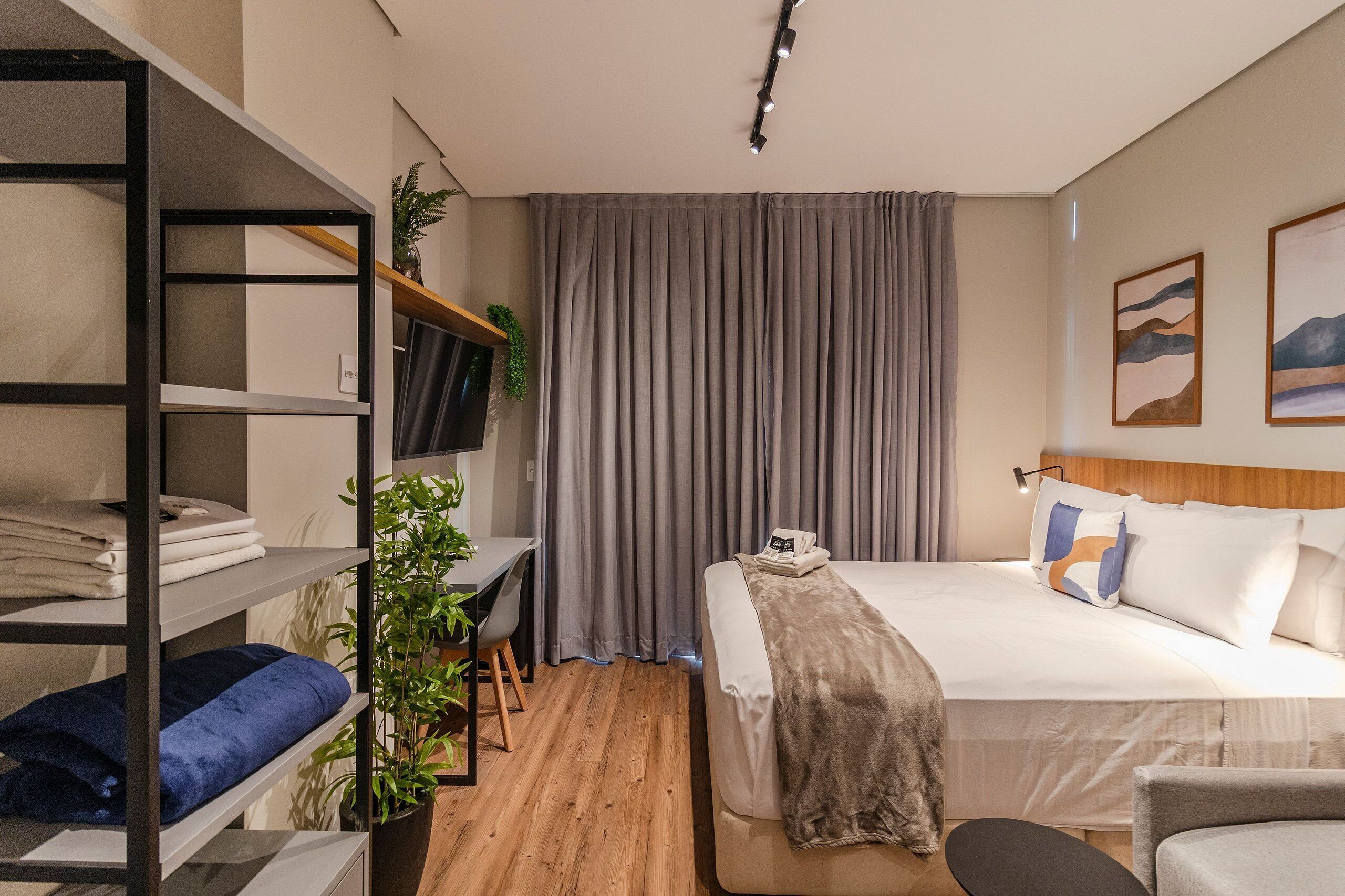 Una habitación de hotel moderna con una cama bien hecha, un escritorio con silla, una estantería y un amplio ventanal con cortinas.