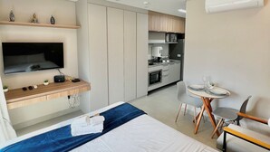 A hotel room with a bed, a desk with a flat-screen TV, a kitchenette, and a dining area with chairs. - La Vida (São Paulo)