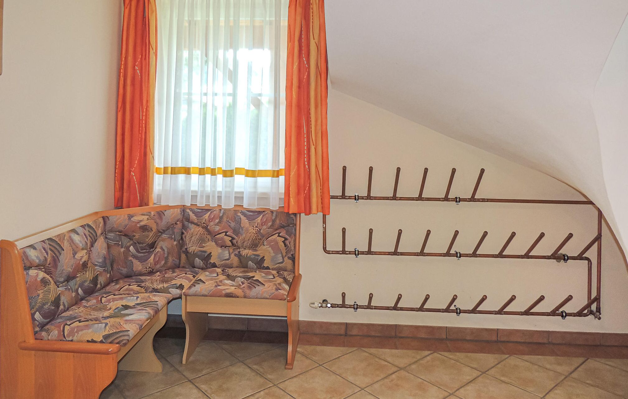 A room with a wooden bench, a window with orange curtains, and a wall-mounted drying rack.