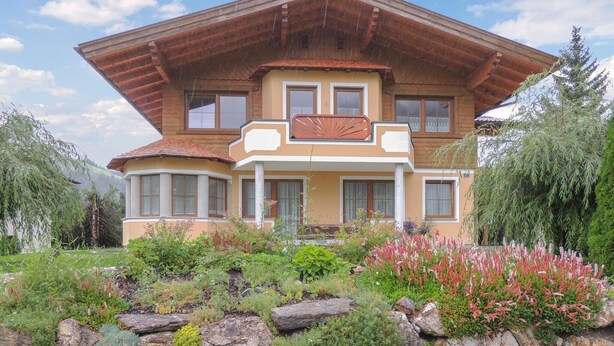 Une maison à deux étages dotée d’un grand balcon, entourée d’un jardin où poussent différentes plantes et fleurs.