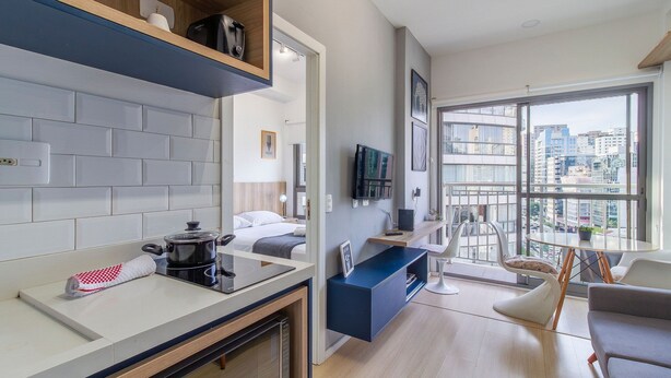 Cocina moderna con encimera, estufa y fregadero. Se observa una zona de comedor con mesa y sillas. Se ve una cama al fondo.