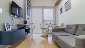 Una sala de estar moderna con un televisor de pantalla plana, un sofá gris, una silla blanca y una mesita con una lámpara.
