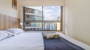 Habitación de hotel con cama, ventana con vistas a edificios altos, un cuadro enmarcado en la pared y una lámpara de noche.